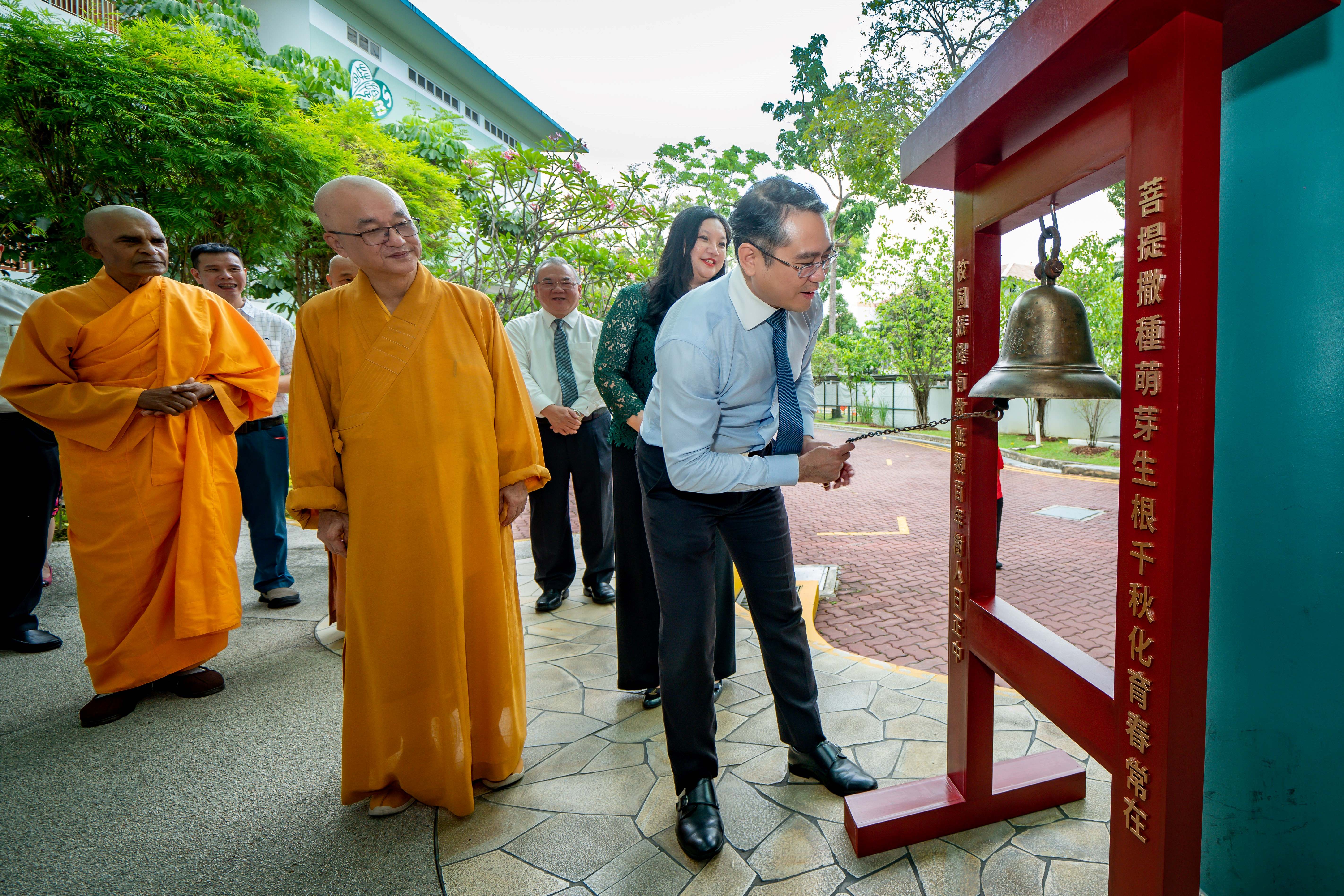 A man in a shirt and tie rings a bronze bell on a red stand with Chinese characters, watched by Buddhist monks in orange robes.